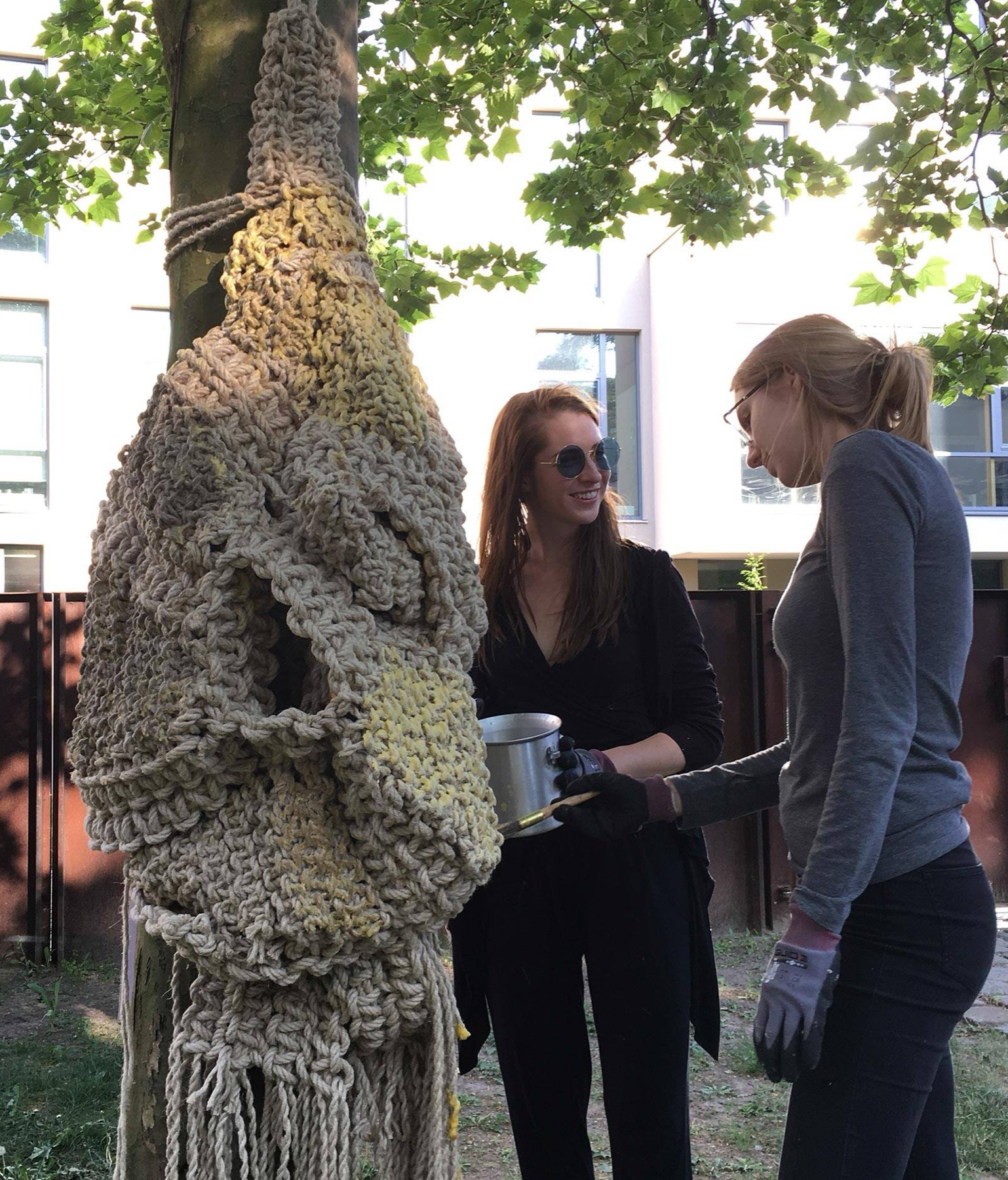 A crocheted rope sleeve tied around a plane tree, two people working on it in the courtyard