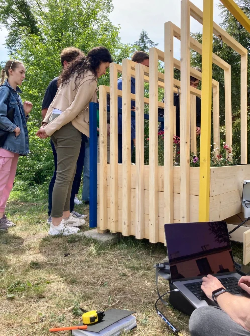 A timber frame going up outdoors, four people working on it, a laptop running on the grass beside them